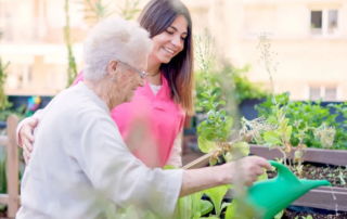 Smiling nurse and senior woman watering plants in a garden.